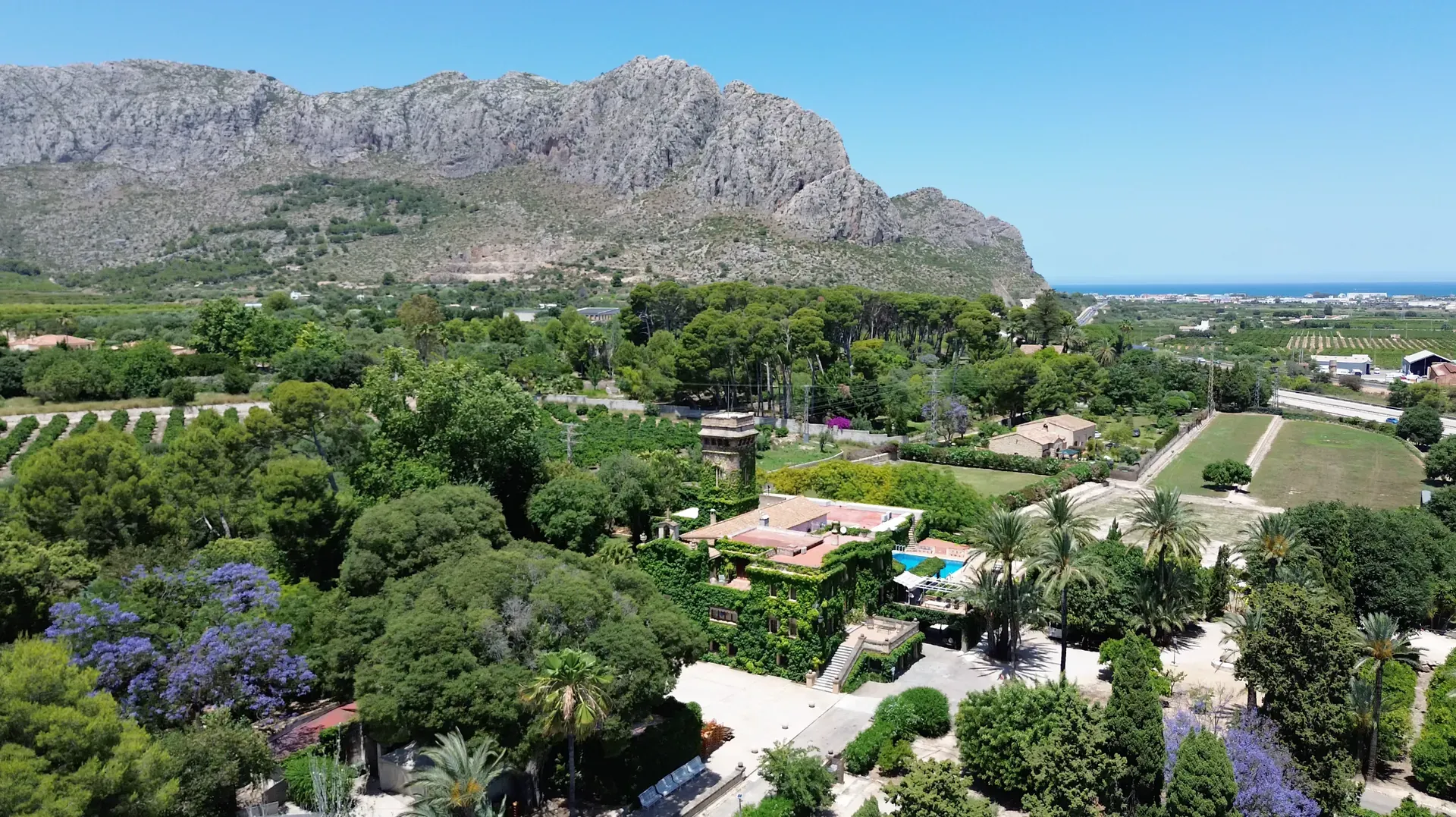Vista aérea de una finca rodeada de jardines y palmeras a los pies de la montaña